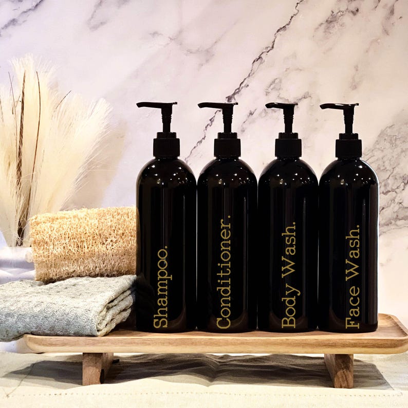 May include: Four black pump bottles with gold labels on a wooden tray. The labels read "Shampoo", "Conditioner", "Body Wash", and "Face Wash".
