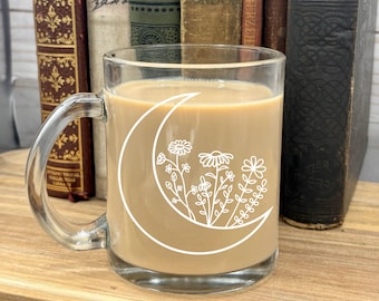 May include: A clear glass mug with a white crescent moon and floral design. The mug is filled with a light brown beverage.