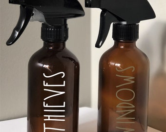 May include: Two amber glass spray bottles with black spray tops. The bottle on the left is labeled "THIEVES" in white lettering. The bottle on the right is labeled "WINDOWS" in gold lettering.