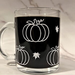 Puede incluir: Una taza de caf&eacute; de vidrio negro con un contorno blanco de calabazas y hojas. La taza est&aacute; llena de caf&eacute;.