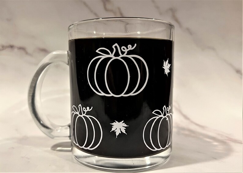 Puede incluir: Una taza de caf&eacute; de vidrio negro con un contorno blanco de calabazas y hojas. La taza est&aacute; llena de caf&eacute;.
