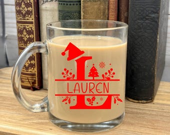May include: A clear glass mug with a red and green Christmas design. The mug has the letter "L" with a Santa hat on top and a Christmas tree next to it. The name "Lauren" is written below the letter. The mug is filled with a light brown beverage.