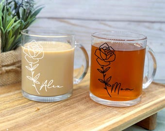 May include: Two clear glass mugs with handles. One mug has a white outline of a rose and the name "Alex" printed on it. The other mug has a black outline of a rose and the name "Mom" printed on it.