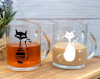 May include: Two glass mugs with handles, one with a black and white striped cat design and the other with a white cat design with stars. Both mugs are filled with a light brown liquid.