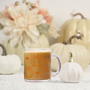 Puede incluir: Una taza de vidrio transparente con asa, llena de un l&iacute;quido marr&oacute;n. La taza tiene un dise&ntilde;o de calabaza naranja con hojas. La taza est&aacute; sobre una superficie blanca con calabazas blancas en el fondo.