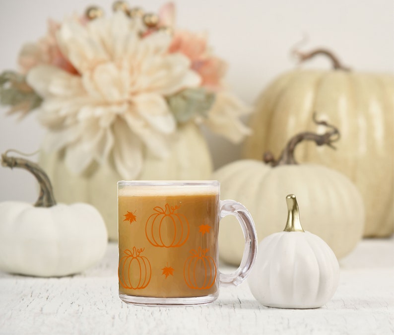Puede incluir: Una taza de vidrio transparente con asa, llena de un l&iacute;quido marr&oacute;n. La taza tiene un dise&ntilde;o de calabaza naranja con hojas. La taza est&aacute; sobre una superficie blanca con calabazas blancas en el fondo.