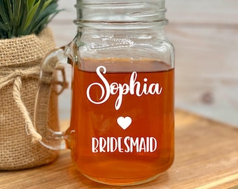 May include: A clear glass mason jar mug with a handle, filled with a dark liquid. The mug has a white vinyl decal with the text "Sophia" in cursive, a small heart, and the word "Bridesmaid".