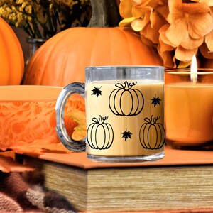 Puede incluir: Una taza de vidrio transparente con un contorno negro de calabazas y hojas. La taza est&aacute; llena de un l&iacute;quido marr&oacute;n claro y est&aacute; sentada sobre una pila de libros. Hay calabazas y una vela encendida en el fondo.