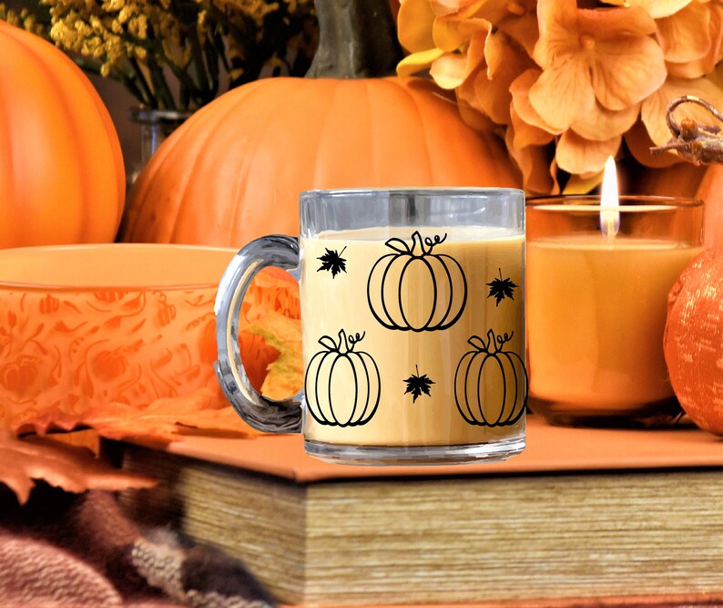 Puede incluir: Una taza de vidrio transparente con un contorno negro de calabazas y hojas. La taza est&aacute; llena de un l&iacute;quido marr&oacute;n claro y est&aacute; sentada sobre una pila de libros. Hay calabazas y una vela encendida en el fondo.