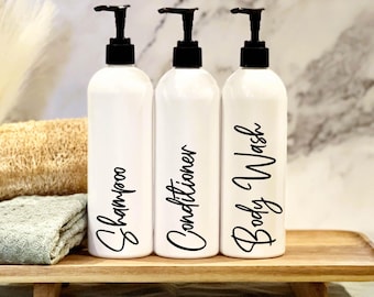 May include: Three white pump bottles with black labels and black pumps. The labels read "Shampoo", "Conditioner", and "Body Wash". The bottles are sitting on a wooden tray.