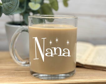 May include: A clear glass mug with a handle, filled with a light brown beverage. The mug has the word "Nana" written on it in white with three stars above the word.