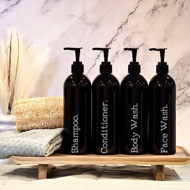 May include: Four black pump bottles with white labels on a wooden tray. The labels read "Shampoo", "Conditioner", "Body Wash", and "Face Wash".