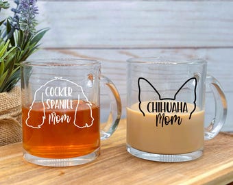 May include: Two ceramic coffee mugs with black handles. The mug on the left has the text "Cocker Spaniel Mom" and a white outline of a cocker spaniel. The mug on the right has the text "Chihuahua Mom" and a black outline of a chihuahua. Both mugs have brown liquid inside.