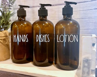 May include: Three amber glass pump bottles labeled "HANDS", "DISHES", and "LOTION". Each bottle has a black pump dispenser. The bottles are arranged on a white wooden tray, suggesting a bathroom or kitchen organization theme.
