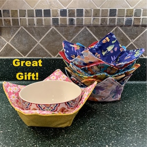 May include: A stack of colorful fabric bowl cozies with floral and patterned designs. One cozy holds a white bowl with a decorative rim. The text "Great Gift!" is in the image. These cozies are designed to keep bowls warm.