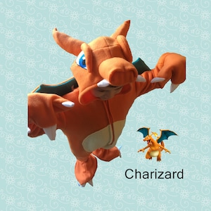 Op de afbeelding: Een kind dat een oranje en groen Charizard-kostuum draagt. Het kostuum heeft een groot hoofd met een puntige snuit en een lange staart. Het kind staat op een witte achtergrond met een bloemenpatroon.