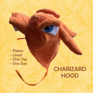 Puede incluir: Una capucha Charizard de forro polar naranja con un ojo azul y blanco, lazos en la barbilla y un interior forrado. La capucha presenta un diseño inspirado en el popular personaje. El texto "CHARIZARD HOOD" se muestra en marrón.