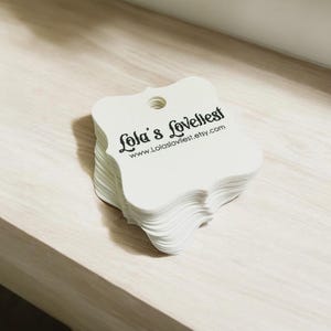 May include: A stack of white, uniquely shaped tags with a small hole at the top. The tags have a scalloped edge and feature the text "Lola's Loveliest" and a website address in black font. The tags are stacked on a light wood surface.