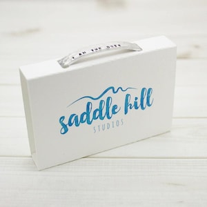 May include: A white box with a clear handle and the text "I AM THE BOSS" printed on the handle. The box features the blue text "saddle hill STUDIOS" on the front. The box is on a white wooden surface.
