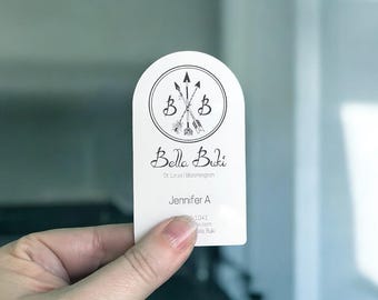 May include: A white business card with a rounded top is held by a hand. The card features a circular logo with arrows and the text "Bella Buki" in script. Additional text includes "St. Louis | Bloomington" and a name.