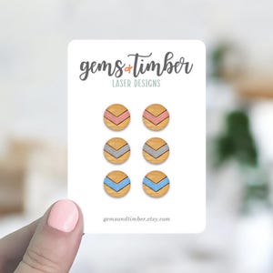 May include: A set of six wooden stud earrings with chevron designs in pink, gray, and blue. The earrings are on a white card with the text "gems + timber LASER DESIGNS" and the website address.