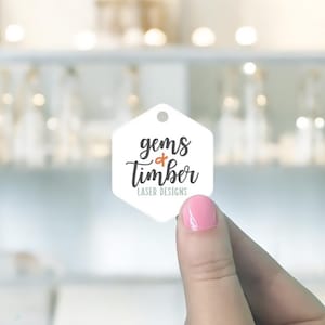 May include: A white, hexagonal tag with a small hole at the top is held up by a person. The tag features the words "gems & timber" in a handwritten font, with "LASER DESIGNS" below. The background is blurred.