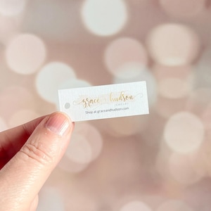 May include: A small, rectangular white tag with the gold text "grace hudson jewelry" and "Shop at graceandhudson.com". The tag has a small hole at the top left corner. The background is a soft, blurred bokeh of pink and white.