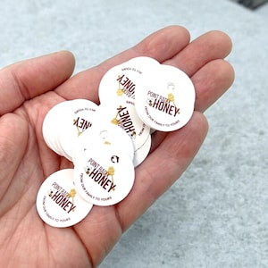 May include: A stack of white, circular tags with a small hole at the top. Each tag features the text "Point Radik Honey" in a brown font, along with a bee graphic. The phrase "From our family to yours" is also printed on each tag.