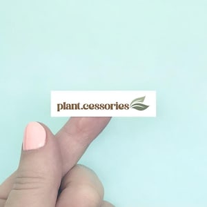 May include: A white rectangular sticker with the text "plant.cessories" in a brown font, and a leaf logo. The sticker is held up by a finger with pink nail polish against a light blue background.