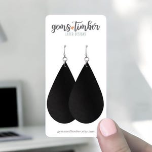 May include: Black teardrop earrings with silver-colored hooks are displayed on a white card. The card has the text "gems+timber LASER DESIGNS" at the top and "gemsandtimber.etsy.com" at the bottom. The earrings are held by a person.