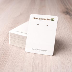 May include: A stack of white earring display cards with rounded corners. One card is upright, displaying two earring holes and the text "plant.cessories" with a leaf logo and the website address.