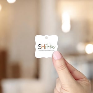 May include: A white, uniquely shaped tag with the text "SHStudios" in a mix of colors. The tag is held by a person with a blurred background. The website address saddlehillstudiosetsy.com is also visible.