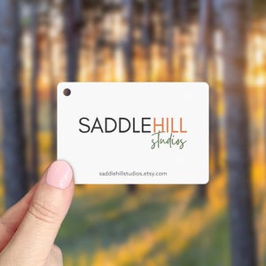 May include: A white rectangular tag with a small hole at the top is held by a person. The tag features the text "SADDLEHILL Studios" in black, orange, and green. The website address saddlehillstudios.etsy.com is printed at the bottom.