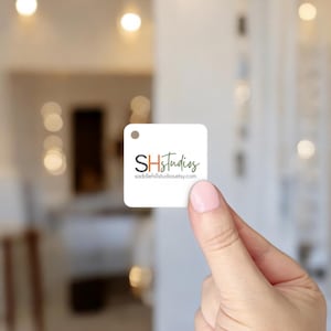May include: A square white tag with a small hole at the top is held by a person. The tag features the text "SHstudios" in black and orange, with the website address in green below.