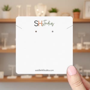 May include: A white earring display card with the logo "SHStudios" in black, orange, and green. The card has two pre-drilled holes for earrings and the website address saddlehillstudios.com. The card is held in front of a wooden shelf with jewelry displays.