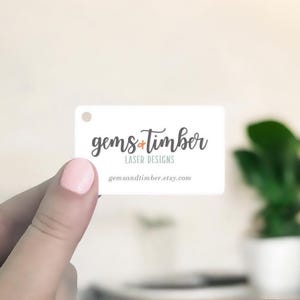 May include: A white rectangular tag with rounded corners, held by a hand. The tag features the text "gems + timber LASER DESIGNS" in a script font, with the Etsy shop URL below. A small hole is at the top.