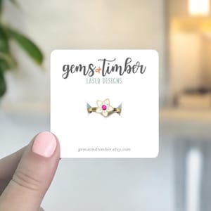 May include: A gold-colored ring with a flower design and a pink center stone is displayed on a white card. The card has the text "gems + timber LASER DESIGNS" and the website address. The ring is held by a person.