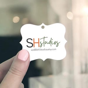 May include: A white, scalloped-edge tag with the text "SHStudios" in black, orange, and green script. The website address saddlehillstudios.etsy.com is below the logo. The tag is held up by a person with pink nail polish.