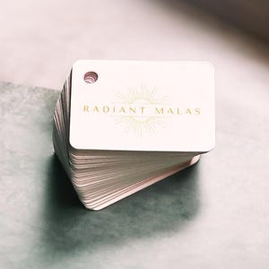 May include: A pile of white, rounded-corner cards featuring a gold logo that reads "RADIANT MALAS" and a sunburst design. Each card has a small hole at the top. The cards are stacked on a grey surface.