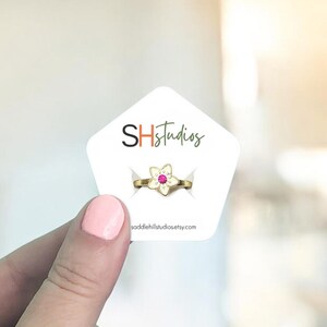 May include: A gold-colored ring with a flower design and a pink gemstone center. The ring is displayed on a white card with the text "SHStudios" and the website "saddlehillstudiosetsy.com".
