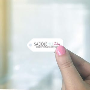 May include: A white tag-shaped sticker with the text "SADDLEHILL studios" and "saddlehillstudios.etsy.com" in black and pink. The sticker is held between fingers with pink nail polish.