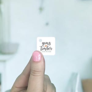 May include: A small, square white tag with the text "gems of timber laser designs" in a handwritten style. The tag is held between fingers with pink nail polish. A small hole is at the top left corner of the tag.