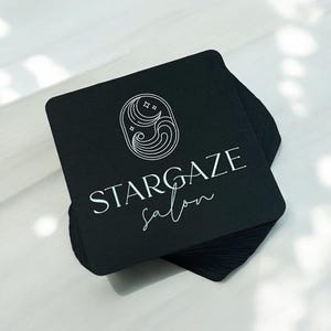 May include: A stack of square black business cards with the text "STARGAZE salon" in white script. The logo features a stylized design of stars and waves. The cards are on a light-colored surface.