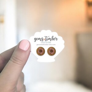 May include: Wooden stud earrings with a floral design. The earrings are round with a brown base and a pink flower. The earrings are displayed on a white scalloped card with the text "gems & timber laser designs".