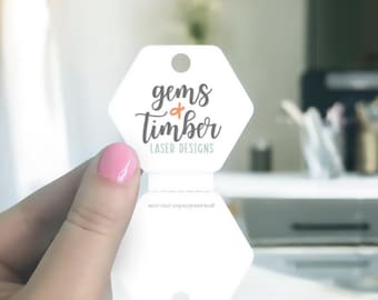 May include: A white, hexagonal tag featuring the text "gems & Timber LASER DESIGNS" in black script and a small orange ampersand. The tag is held by a person with pink nail polish.