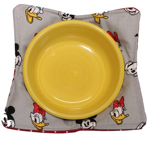 May include: A gray and red fabric bowl mat with a white polka dot border. The mat features a repeating pattern of cartoon characters, including Mickey Mouse and Minnie Mouse. A yellow ceramic bowl sits on top of the mat.