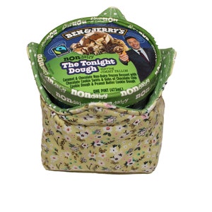 May include: A green and yellow fabric ice cream container with a Ben & Jerry's pint of The Tonight Dough non-dairy ice cream inside. The ice cream container has a picture of Jimmy Fallon on it. The text on the container reads "nondairy The Tonight Dough starring JIMMY FALLON Caramel & Chocolate Non-Dairy Frozen Dessert with Chocolate Cookie Swirls & Gobs of Chocolate Chip Cookie Dough & Peanut Butter Cookie Dough ONE PINT (473ml)"
