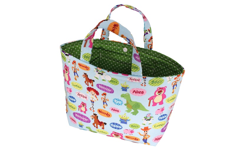 K&ouml;nnte beinhalten: Eine hellblaue Tragetasche mit gr&uuml;nem Innenfutter mit wei&szlig;en Punkten. Die Tasche hat ein sich wiederholendes Muster mit Toy Story-Charakteren, darunter Woody, Buzz Lightyear, Jessie und Lotso. Die Tasche hat zwei Stoffgriffe.