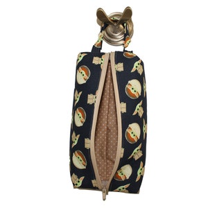 Può includere: Una pochette nera con cerniera con un motivo ripetuto di facce di Baby Yoda dei cartoni animati. La pochette ha una cerniera marrone e una fodera in tessuto marrone con pois bianchi. La pochette è appesa a un gancio dorato.