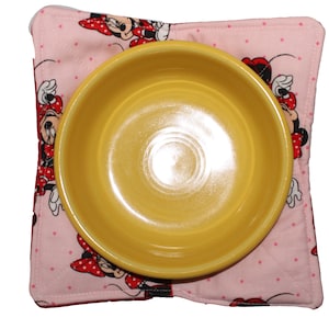 May include: A yellow ceramic bowl sits on a pink and white fabric placemat with a Minnie Mouse design. The placemat has white polka dots and features Minnie Mouse in various poses.